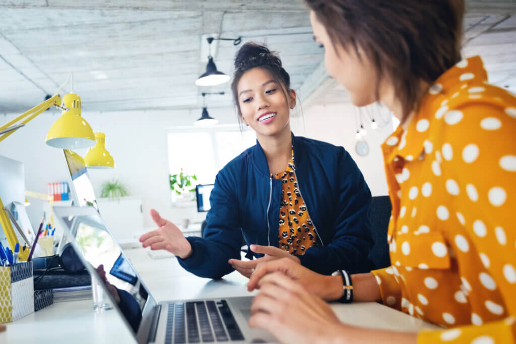 Why is onboarding important? Your secret weapon for recruiting top talent. CJ Talent. Image of two women at a desk, dressed in business casual clothing, talking, with a laptop between them.