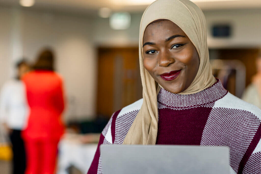 female part time finance director, woman of colour wearing a hijab headscarf, smiles at camera and holds a digital tablet.